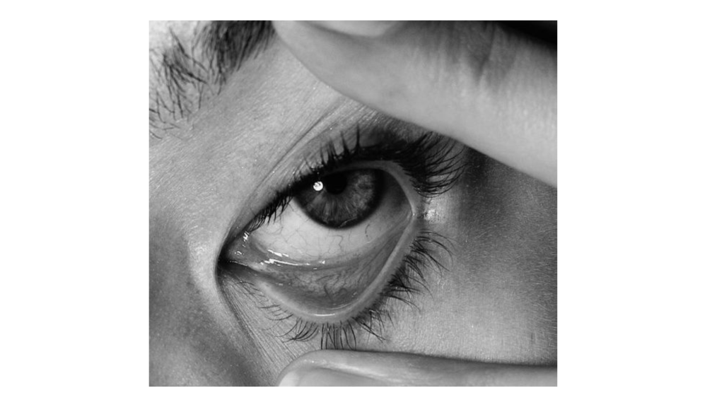 Un primer plano en blanco y negro de un ojo humano, con los dedos tirando suavemente hacia abajo del párpado inferior, revela detalles que reflejan profundidad, del mismo modo que Consultoría de Transformación Cultural y Liderazgo Consciente descubre nuevas perspectivas.