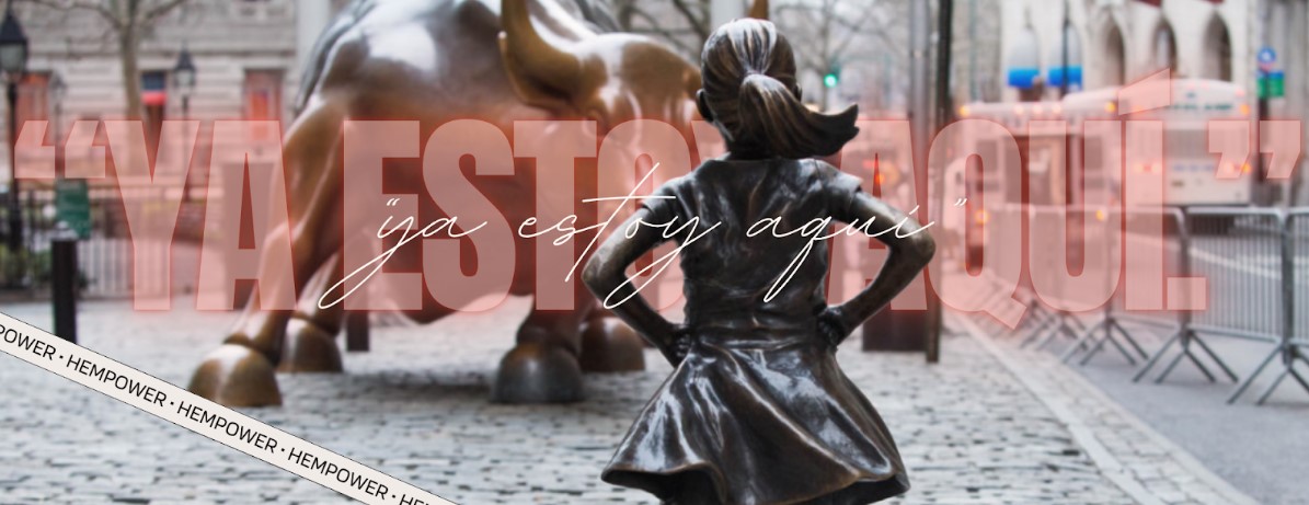 Una estatua de bronce de una chica se enfrenta confiada a la estatua del Toro Bravo en una calle adoquinada. Un texto superpuesto en español dice "YA ESTOY AQUÍ" y "ya estoy aquí". La escena se sitúa en una zona urbana, delimitada por barreras.