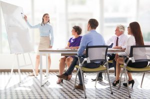 Una mujer está de pie junto a una pizarra dando una presentación sobre Coaching Empresarial para el éxito de tu negocio a cuatro colegas sentados a una mesa. El grupo observa atentamente y la sala está iluminada con luz natural.