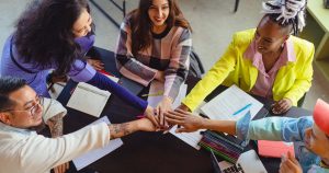 Cinco personas se sientan alrededor de una mesa cubierta de cuadernos, papeles y un ordenador portátil, juntando las manos en una muestra de trabajo en equipo y unidad: un momento inspirador que encarna el Coaching Empresarial para el éxito de tu negocio.